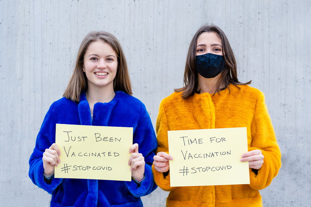 Picture of two women. One is not wearing a mask and holding a sign that says " Just Been Vaccinated. #Stopcovid. The other is wearing a mask and holding a sign that says "Time for vaccination. #stopcovid."